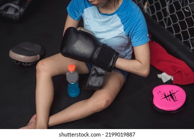 A Novice Female MMA Practitioner Removes Her Boxing Gloves While Sitting Down On The Mat, Exhausted.