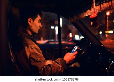 NOVOSIBIRSK, RUSSIA - 20 February 2019: A Man Sits In A Car Cab At Night And Looks At The Phone. Light Of Night Spotlights. The Driver Is Distracted By The Phone While Driving.