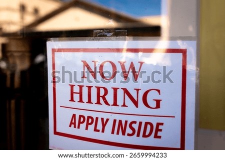 Now Hiring Apply Inside window sign at a local store, a laminated white paper sign with red text, taped to the window of the storefront