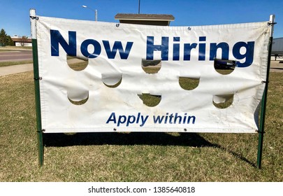 A Now Hiring, Apply Within Sign Appears On A Lawn Against A Blue Sky.