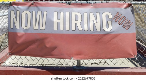 NOW HIRING Help Wanted Banner Hanging On Fence.