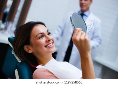 Now my smile is perfect! Beautiful young woman looking at mirror with smile in dentist’s office

