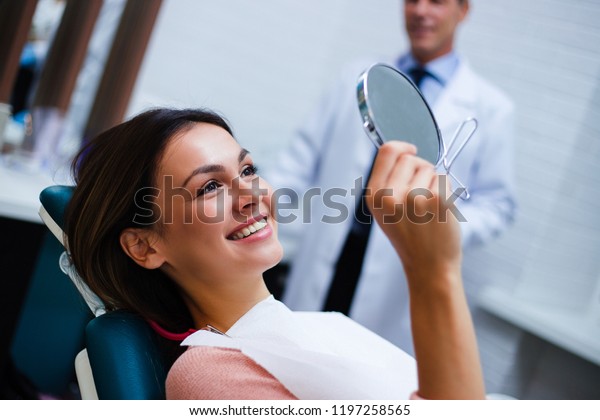 Now my smile is perfect!\
Beautiful young woman looking at mirror with smile in dentist’s\
office\
