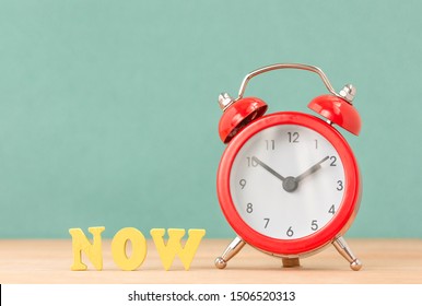 NOW Word Near Alarm Clock On Wooden Desk. Concept Of Present Moment. Time Take Action.