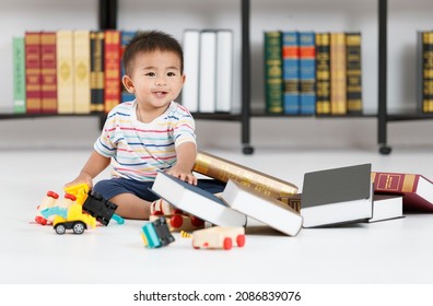 Nuaghty And Cheerful Asian Kid On Casual Lovely Shirt Playing Fun With Toy Car And Reaching Out To Pile Of Fake Books. Little Child Enjoy Messing Up On Library Floor Of Preschool.