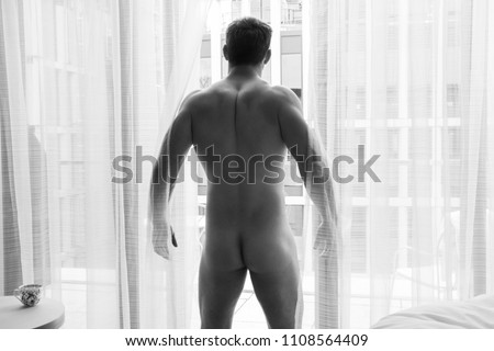 Nude good looking man shows his backside and butt, bottom, as he stands in front of curtains of the balcony of his hotel room