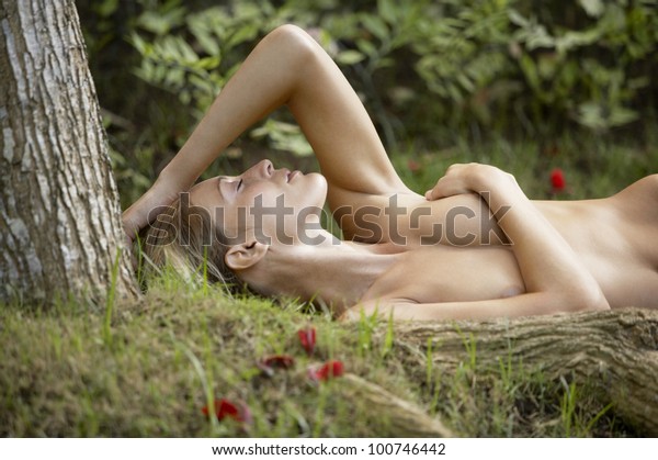 Nude young woman laying down on green grass and red rose petals, with tree roots in the foreground.