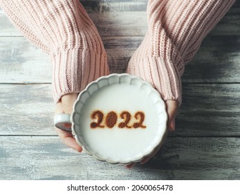 Number 2022 On Frothy Surface Of Cappuccino Served In Pastel Pink Cup Holding By Female Hands Over Grey Painted Wood Planks Table. Holidays Food Art Theme Happy New Year 2022, New Year New You.