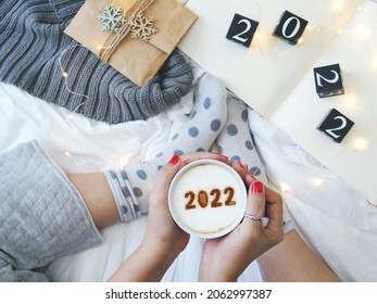 Number 2022 on frothy surface of cappuccino served in white cup by female hands on the bed with white blanket, black painted wooden dice with number 2022, gift bag, knitted scarf. (selective focus)