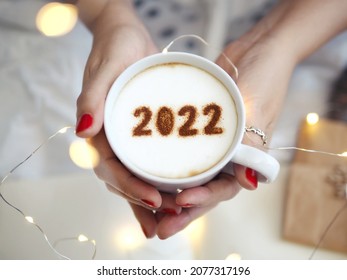 Number 2022 on frothy surface of cappuccino served in white cup holding by female hands on blurred white bed with string lights. New year new you, Holidays food art theme Happy New Year. (top view)