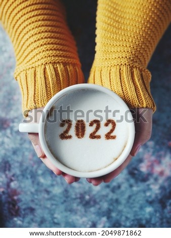 Number 2022 on frothy surface of cappuccino served in white cup holding by female hands over rustic blue background. Holidays food art theme Happy New Year 2022, New year new you. (selective focus)