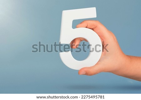 Number five in hand. Hand holding white number 5 on blue background with copy space. Concept with number five. Birthday 5 years, fifth grade, five day work week