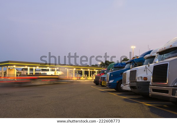 Number of semi trucks of various models and colors\
in the future on the night truck stop with a lit gas station and\
blurred lights of a passing semi truck and reflection of the lights\
on the big rigs.