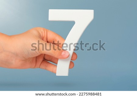 Number seven in hand. Hand holding white number 7 on blue background with copy space. Concept with number seven. Birthday 7 years, percent, seventh grade or day.