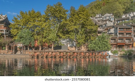 A number of tourist boats with lifebuoys and awnings are moored in a row near the riverbank. Reflection on the water. Ancient Chinese buildings with curved roofs on the coast among green trees. China. - Powered by Shutterstock