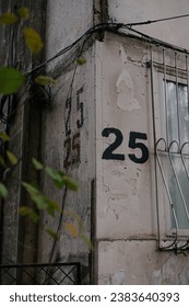 The number twenty five is painted using a stencil three time on a gray wall on the corner of the house, near window with bars. House numbering 25. 