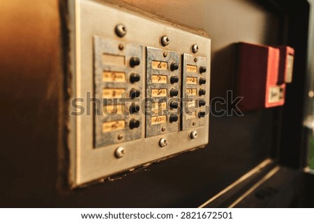 A numbered apartment intercom panel sits beside a red fire alarm pull station on a dark building entrance wall. Warm side light reveals the worn metal textures.