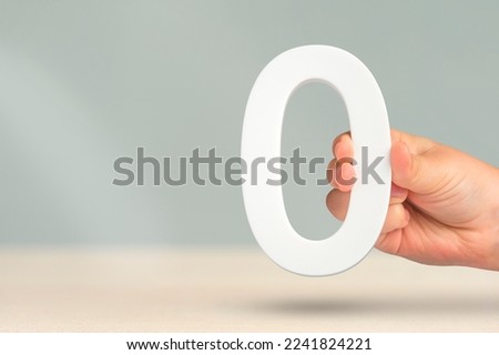 Numeral zero in hand. A hand holds a white number zero on a blurred background with copy space. Zero concept, 0 percent interest rate, minimum air emissions, cost or credit no increase.