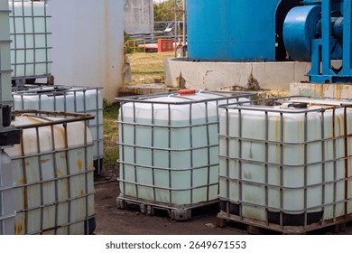 Numerous intermediate bulk containers (IBCs) with metal cages storing liquids in an industrial setting, with large tanks and machinery in the background.