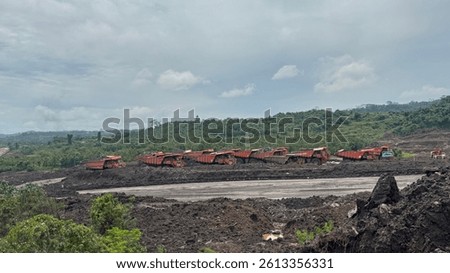 Numerous large mining trucks, laden with coal, are parked in an openpit mine. The backdrop is a verdant hillside under a cloudy sky. Industrial mining landscape.