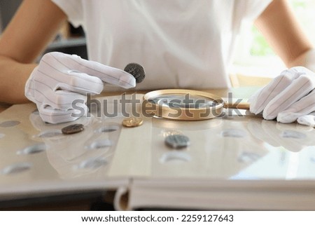 Numismatist in white gloves examines ancient coins collection at table. Close-up of woman with magnifying glass values coins.