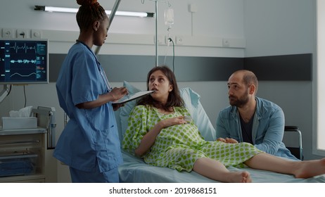 Nurse Of African American Ethnicity Talking To Pregnant Patient While Using Tablet For Pregnancy Healthcare In Hospital Ward. Expecting Husband And Wife Waiting For Child Delivery