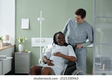 Nurse assisting pregnant woman in wheelchair, both smiling in hospital room with medical equipment and neatly organized space visible - Powered by Shutterstock