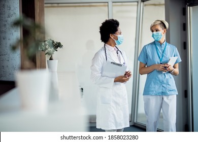 Nurse And Black Female Doctor With Protective Face Masks Talking While Walking In A Hallway At Medical Clinic. 