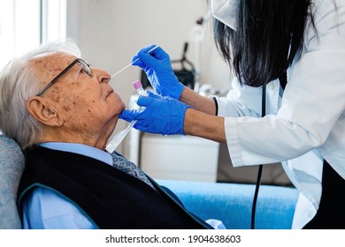 Nurse In Blue Sanitary Gloves Lifts The Head Of An Elderly Man In Blue Shirt And Tie To Perform The Covid Test While He Is Sitting On The Couch At Home. Home Health Care.