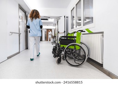 Nurse in blue scrubs walking away in a bright hospital hallway, with an empty green wheelchair on the side. - Powered by Shutterstock