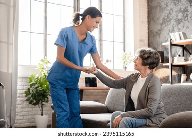Nurse caretaker caregiver doctor helping assisting aiding old elderly disable immobile handicapped patient grandmother stand up from the couch. Rehabilitation after injury trauma - Shutterstock ID 2395115165