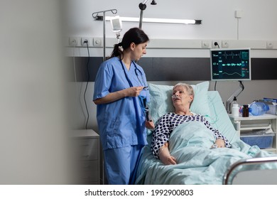 Nurse Discussing With Elderly Patient Laying In Hospital Bed During Visit About Chest X-ray. Recieving Treatment Through Intravenous Line And Inhale And Exhale With Help From Oxygen Mask.