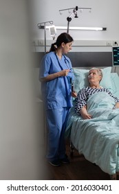 Nurse Discussing With Elderly Patient Laying In Hospital Bed During Visit About Chest X-ray. Recieving Treatment Through Intravenous Line And Inhale And Exhale With Help From Oxygen Mask.