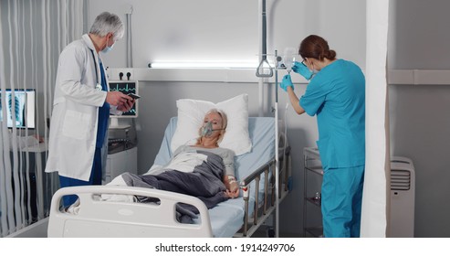 Nurse And Doctor Standing At Hospital Bed And Checking Patient Conditions. Medical Staff Examining Sick Senior Woman In Oxygen Mask Lying In Bed At Hospital Ward