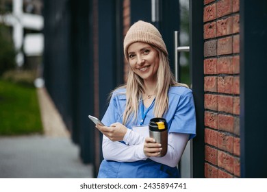 Nurse, doctor in uniform taking break, standing in front hospital building, scrolling on smartphone. Work-life balance of healthcare worker. - Powered by Shutterstock