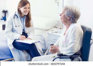 Nurse Or Doctor Writing Patient Notes In A Ward For A Smiling Elderly Woman Seated In A Wheelchair In A Medical And Healthcare Concept