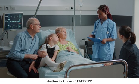 Nurse doing consultation with sick woman while family visits her in hospital ward bed. Medical assistant talking to elder woman and people about illness. Mother, kid and man in visit