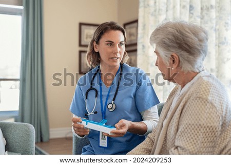 Nurse explaining medicine dosage to old patient in care facility centre while holding weekly medicine dispenser. Caregiver holding pill organizer box giving medicine tablet to elderly woman.