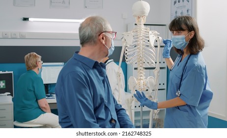 Nurse With Face Mask Explaining Bones On Human Skeleton, Talking About Osteopathy Anatomy And Spinal Cord Pain With Senior Man In Cabinet. Medic Showing Back, Physical Examination.