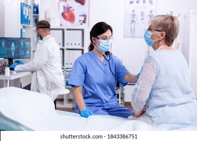 Nurse With Face Mask Listening Patient Heart Using Stethoscope In Hospital Examination Room Wearing Protection Gear. Medical Examination For Infections, Disease And Diagnosis.