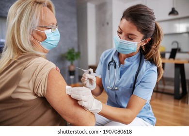 Nurse With Face Mask Sitting At Home With Senior Woman And Injecting Covid 19 Vaccine.