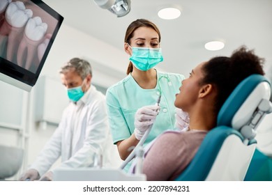 Nurse With Face Mask Using Suction Tube While Preparing African American Woman For Treatment At Dental Clinic.