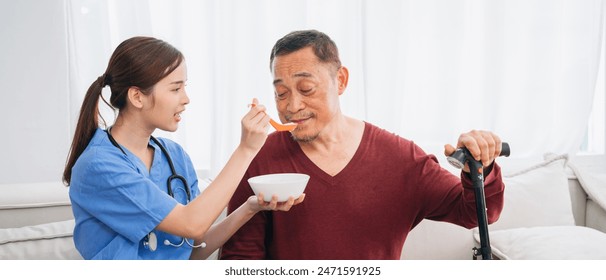 Nurse is feeding a man a bowl of food. The woman is wearing a blue shirt and the man is wearing a red shirt. The man is using a cane to help him stand up - Powered by Shutterstock