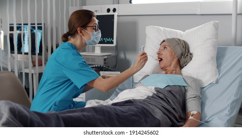 Nurse Feeding Mature Woman Having Cancer With Porridge. Medical Worker In Safety Mask And Scrubs Feeding Ill Senior Female Patient Lying In Bed After Chemotherapy