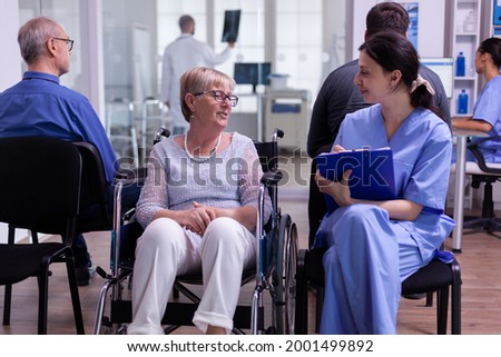 Nurse filing documents while talking with disabled senior woman sitting in wheelchair in hospital waiting area. Elderly paralyzed patient explaning symptoms, assitant checking registration form