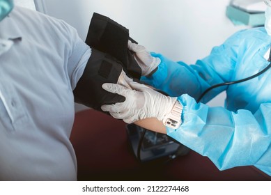 A Nurse In Full PPE Gear Straps On A Inflatable Cuff On A Patient At An ER. Checking Blood Pressure With A Sphygmomanometer