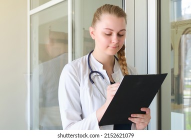 Nurse Girl Doctor On Gray Background Holding A Folder In Her Hands Smiling Writes A Blonde Beautiful. Concept Student Trainee Report Task Attentiveness.