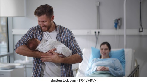 Nurse giving newborn son wrapped in blanket to young father in hospital. Happy man holding baby with wife resting in hospital bed after childbirth on background