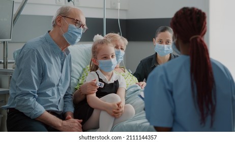 Nurse Giving Recovery Advice To Patient And Visitors With Face Mask In Hospital Ward. Medical Assistant Helping With Medication At Checkup Visit, Talking To Sick Woman And Family During Pandemic.