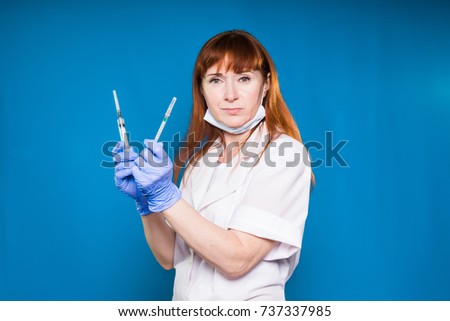 a nurse with gloves holds two syringes in her hands and looks strictly at the camera, isolated on a blue background
